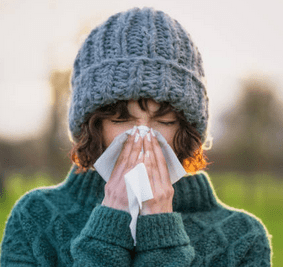 Personal health care outdoor picture of a woman wearing a sweater and a toque sneezing with her two hands placed over her nose in a handkerchief