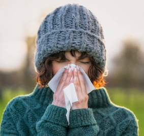 Personal Health Care Picture of a woman wearing a gray wool jumper and a hat, sneezing into a tissue with both hands